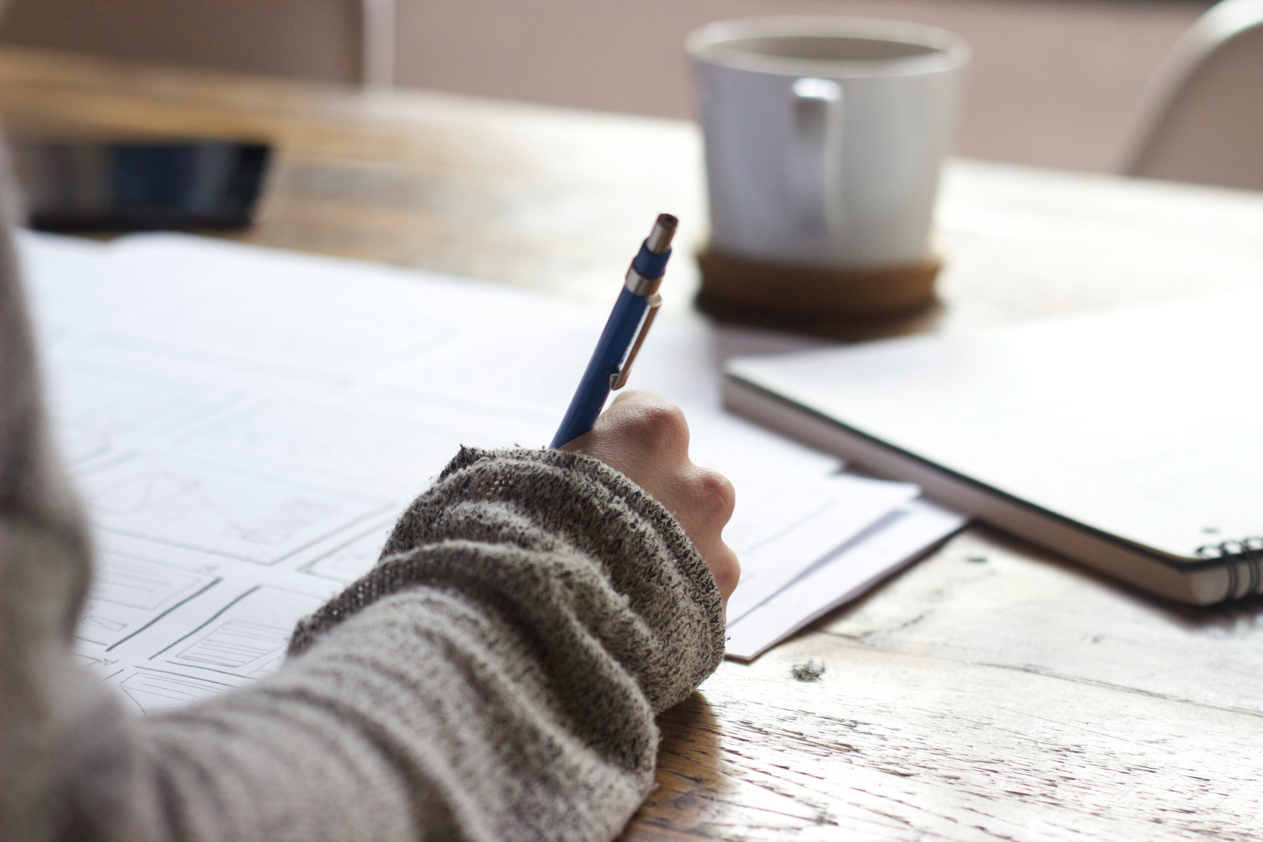A person writes on paper at a table with coffee. 